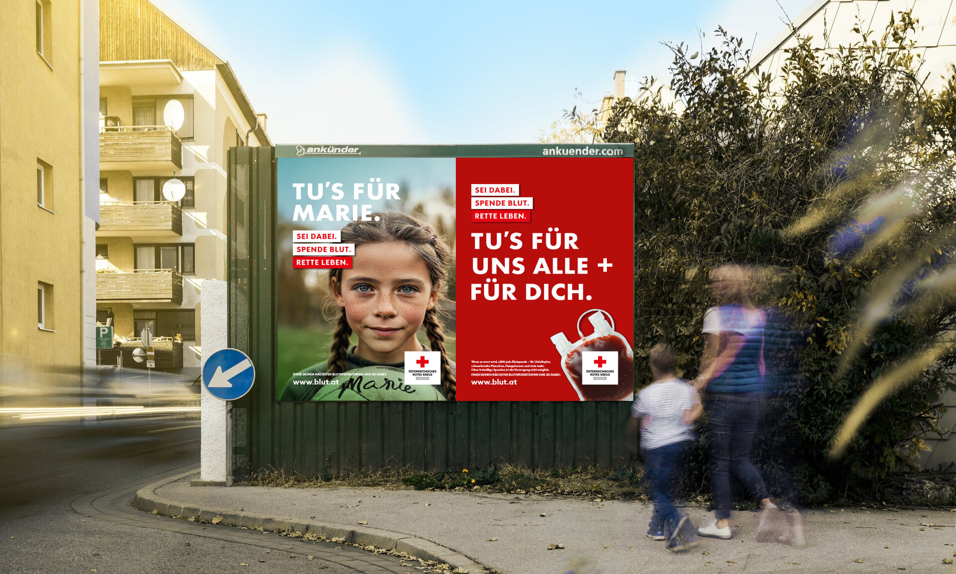 Großes Plakat an einer Straße. Links sieht man ein Bild eines jungen Mädchens mit geflochtenen Zöpfen. Daneben steht weißer Text mit rotem Hintergrund: „Tu’s für Marie. Sei dabei. Spende Blut. Rette Leben.“ Rechts daneben ist ein rotes Plakat mit einem Blutbeutel und dem Text: „Tu’s für uns alle + für dich.“ Vor dem Plakat gehen verschwommen eine erwachsene Person und ein Kind vorbei. Im Hintergrund stehen Häuser und Büsche.