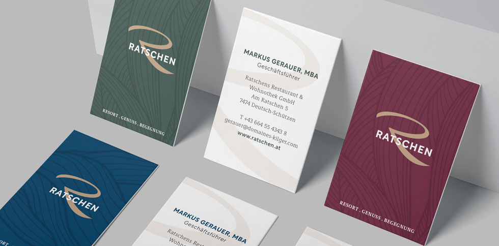 Mehrere Ratschen-Visitenkarten liegen nebeneinander. Die Vorderseiten sind in Grün, Blau oder Weinrot mit Logo gestaltet. Die Rückseiten zeigen Kontaktdaten auf weißem Hintergrund.
