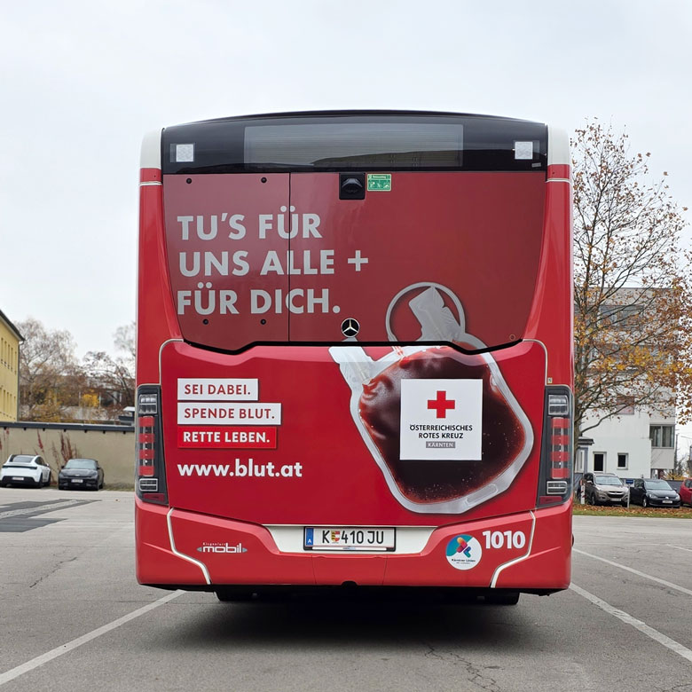 Ein Linienbus von hinten. Die Rückseite ist komplett rot gestaltet. Darauf steht in großen weißen Buchstaben: „Tu’s für uns alle + für dich.“ Unten links stehen die Worte „Sei dabei. Spende Blut. Rette Leben.“ Daneben ist ein Bild eines Blutbeutels mit dem Logo des Österreichischen Roten Kreuzes. Unten steht die Website www.blut.at