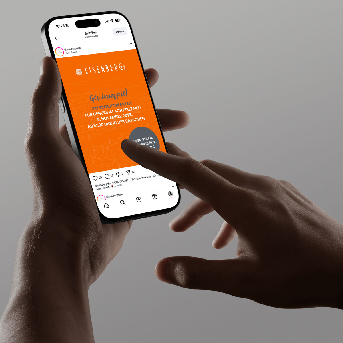 Eine Person hält ein Smartphone in beiden Händen. Auf dem Bildschirm ist ein Instagram-Posting der Eisenberg DAC zu sehen. Der Beitrag hat einen orangefarbenen Hintergrund und kündigt ein Gewinnspiel für „Genuss im Achterltakt“ an. Die Finger der Person berühren den Bildschirm, als würde sie scrollen oder tippen. Der Hintergrund ist grau und unscharf.