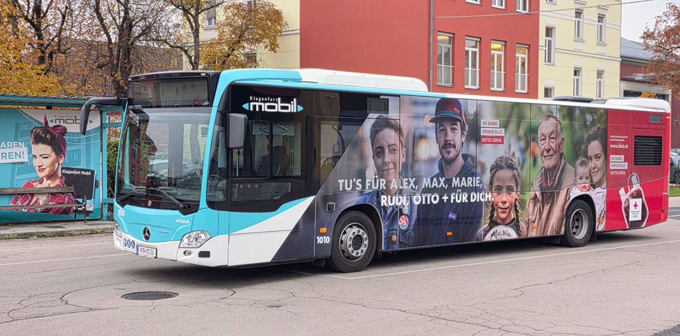 Ein Linienbus von der Seite, teilweise türkis, teilweise rot. Auf der roten Fläche sieht man Fotos verschiedener Menschen: eine Frau mit einem Baby, ein junger Mann mit Kappe, ein Mädchen mit Zöpfen und ein älterer Mann. Der Text lautet: „Tu’s für Alex, Max, Marie, Rudi, Otto + für dich.“ Daneben stehen die Worte: „Sei dabei. Spende Blut. Rette Leben.“ Rechts am Bus ist ein großes Bild eines Blutbeutels mit dem Logo des Österreichischen Roten Kreuzes.