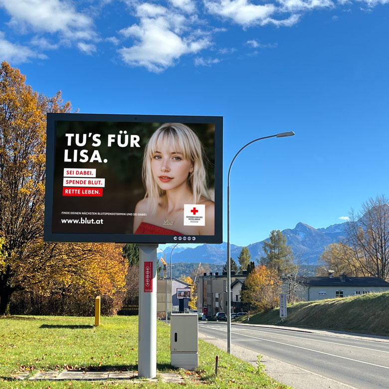 Digitale Werbetafel an einer Straße mit Bergen im Hintergrund. Auf der Anzeige ist eine junge Frau mit blondem Haar zu sehen. Der Text lautet: „Tu’s für Lisa. Sei dabei. Spende Blut. Rette Leben.“ Unten stehen das Logo des Österreichischen Roten Kreuzes und die Website www.blut.at
