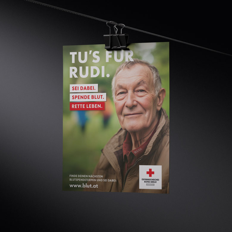 Ein Plakat hängt mit einer Klammer an einer schwarzen Schnur. Darauf sieht man einen älteren Mann, der freundlich in die Kamera blickt. Der Text lautet: „Tu’s für Rudi. Sei dabei. Spende Blut. Rette Leben.“ Unten stehen das Logo des Österreichischen Roten Kreuzes und die Website www.blut.at