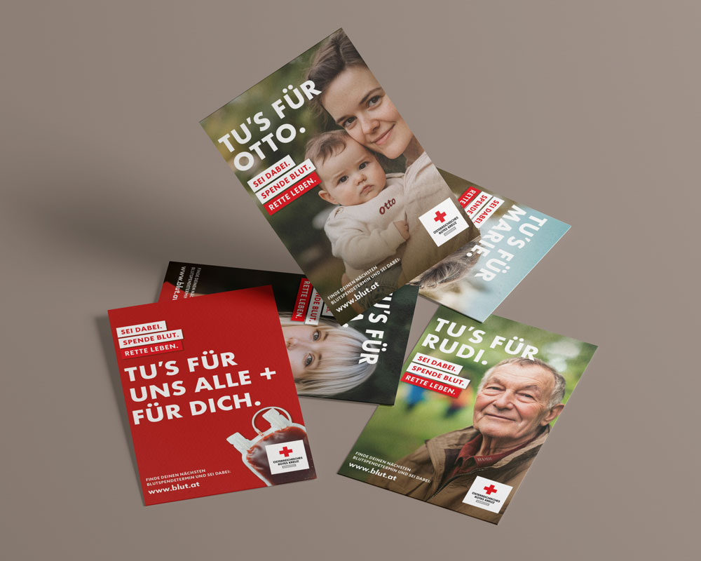 Mehrere Postkarten liegen locker auf einer braunen Fläche. Eine Karte zeigt ein Baby im Arm einer Frau mit dem Text: „Tu’s für Otto. Sei dabei. Spende Blut. Rette Leben.“ Eine weitere Karte zeigt einen älteren Mann mit dem Text: „Tu’s für Rudi.“ Eine dritte Karte zeigt ein rotes Motiv mit einem Blutbeutel und dem Text: „Tu’s für uns alle + für dich.“ Außerdem sind Karten mit weiteren Personen teilweise sichtbar. Alle Karten tragen das Logo des Österreichischen Roten Kreuzes und die Website www.blut.at