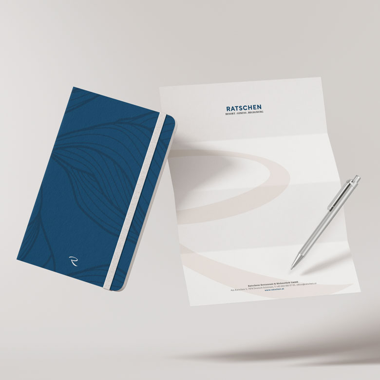 Ein blaues Notizbuch mit feinen Linien liegt neben einem gefalteten weißen Briefpapier der Ratschen. Auf dem Briefpapier steht oben das Ratschen-Logo. Daneben liegt ein silberner Stift.