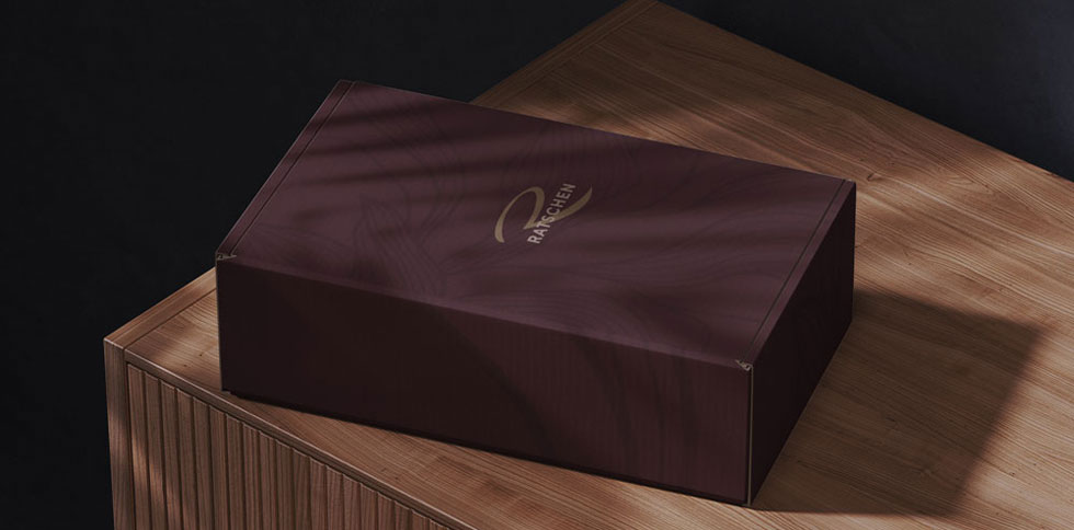 Eine weinrote Geschenkbox mit dem Ratschen-Logo liegt auf einer hellen Holzkommode. Weiches Licht wirft Schatten über die Box.