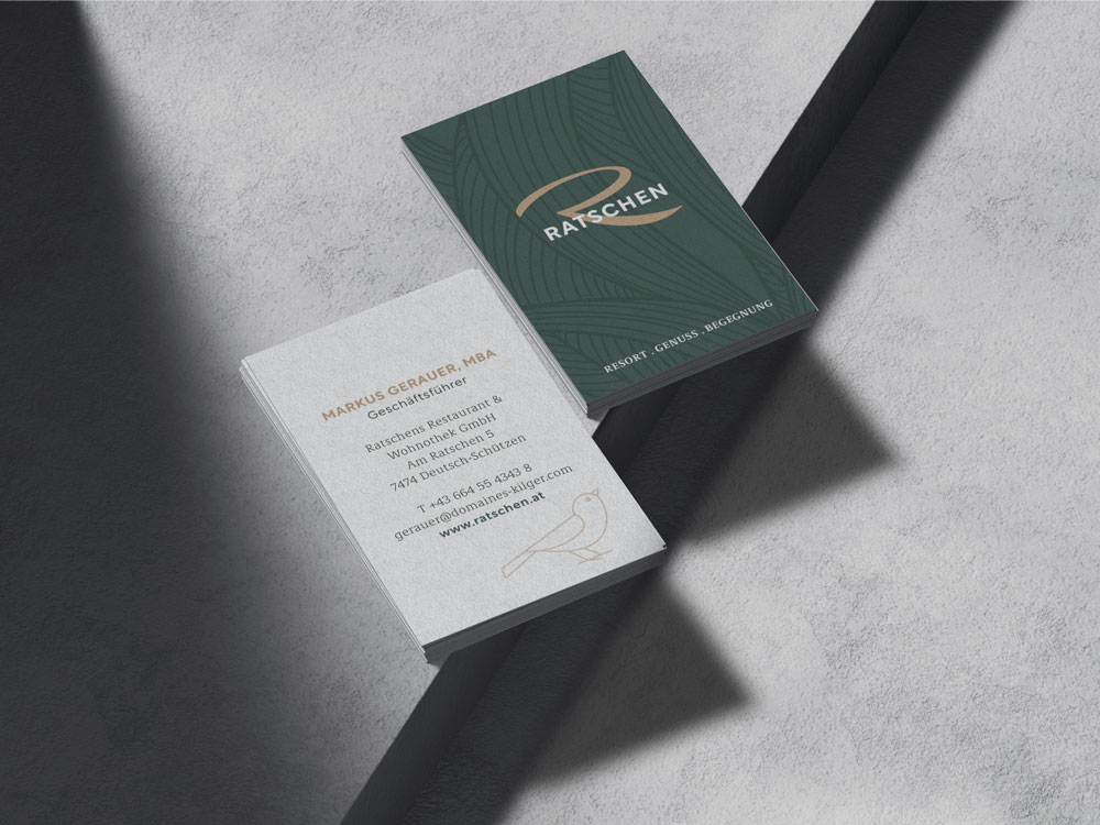 Zwei Stapel Ratschen-Visitenkarten liegen auf einer Steinoberfläche. Eine Karte zeigt Kontaktdaten, die andere das dunkelgrüne Logo-Design. Licht und Schatten fallen über die Karten.