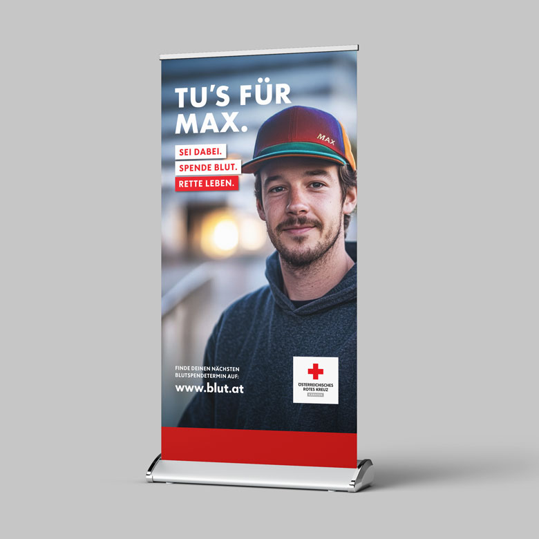 Roll-Up-Banner vor grauem Hintergrund. Darauf ist ein junger Mann mit Kappe zu sehen. Text: „Tu’s für Max. Sei dabei. Spende Blut. Rette Leben.“ Unten steht die Website www.blut.at