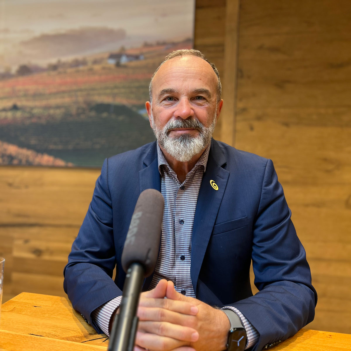 Porträt eines Mannes mit grauem Bart und Anzug, der an einem Holztisch sitzt und in ein Mikrofon spricht – Interview- oder Gesprächssituation in warmem, holzgetäfeltem Raum.